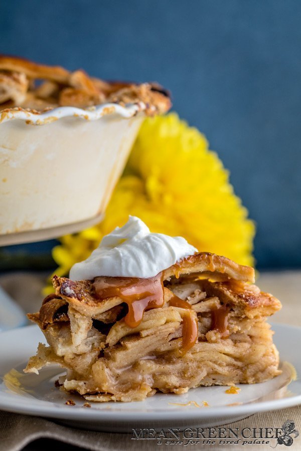 Side photo of a huge slice of Caramel Apple Pie on a white plate garnished with caramel and soft whipped cream. With a pie in the far background. Mean Green Chef