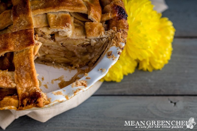 Forty-five degree photo of Caramel Apple Pie Cut into with a slice missing and yellow flowers in the background. Mean Green Chef