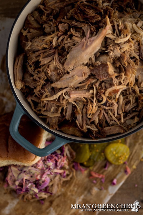 Overhead photo of Slow Roasted Pulled Pork in a gray Dutch Oven with sandwich off the the side in the background. Mean Green Chef