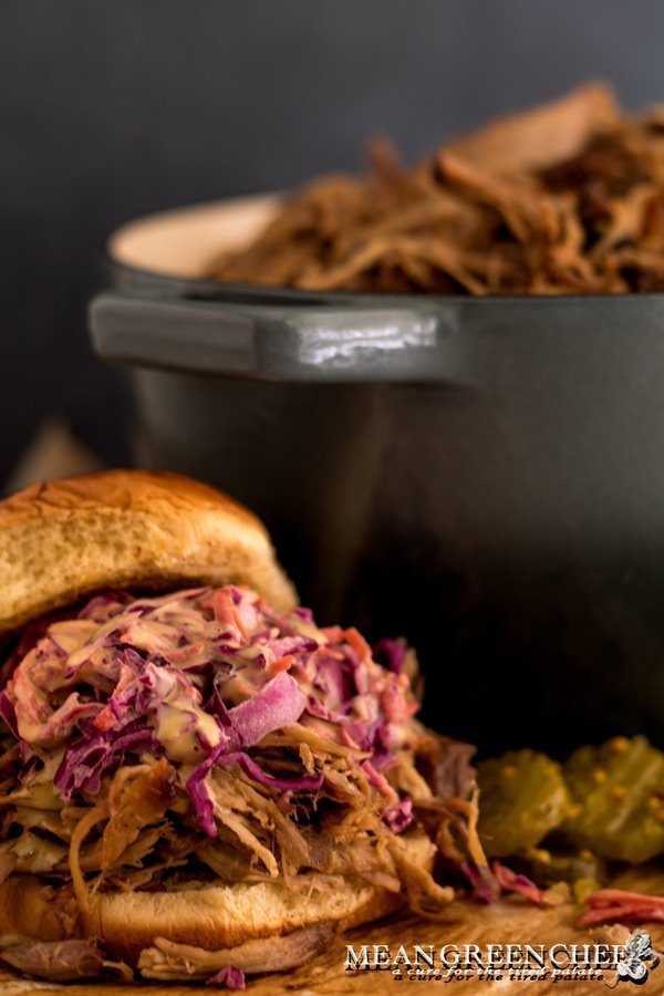 Side photo of a large puled pork sandwich piuiled high with Red Cabbage Carrot Slaw with a gray pot in the background filled with more pulled pork and pickles off to the side of the sandwich. Mean Green Chef
