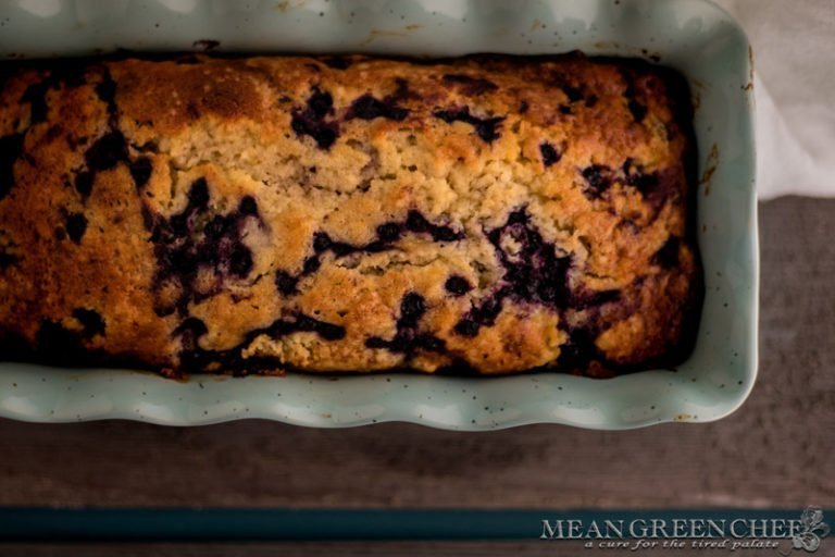 Overhead photo of Rustic Blueberry Banana Bread baked in a blue scalloped loaf pan on a gray wooden background. Mean Green Chef