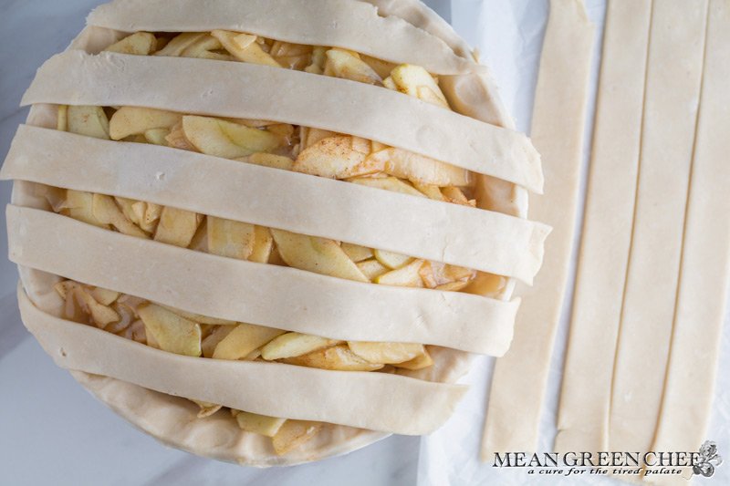 Overhead photo of Lattice Pie Crust being created over a caramel apple pie, photographed on a parchment paper background. Mean Green Chef