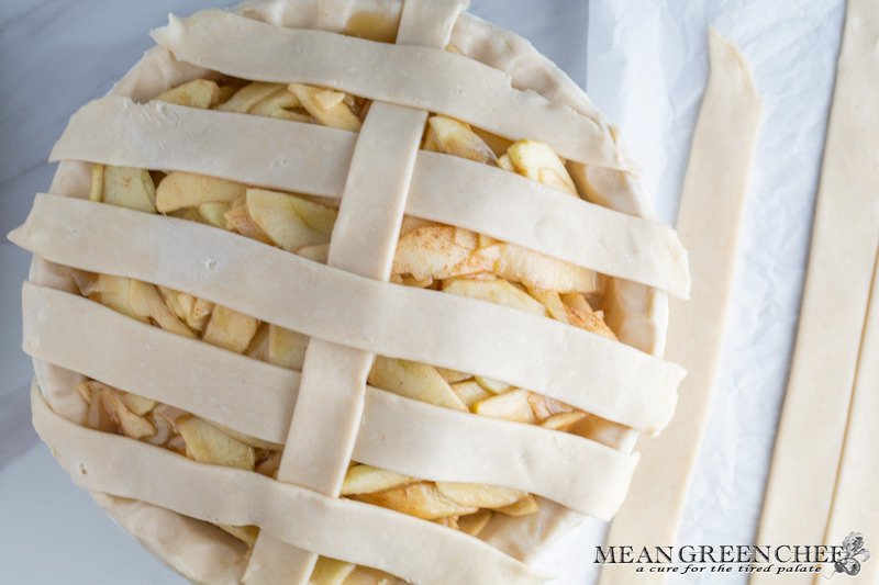 Overhead photo of Lattice Pie Crust being created over a caramel apple pie, photographed on a parchment paper background. Mean Green Chef