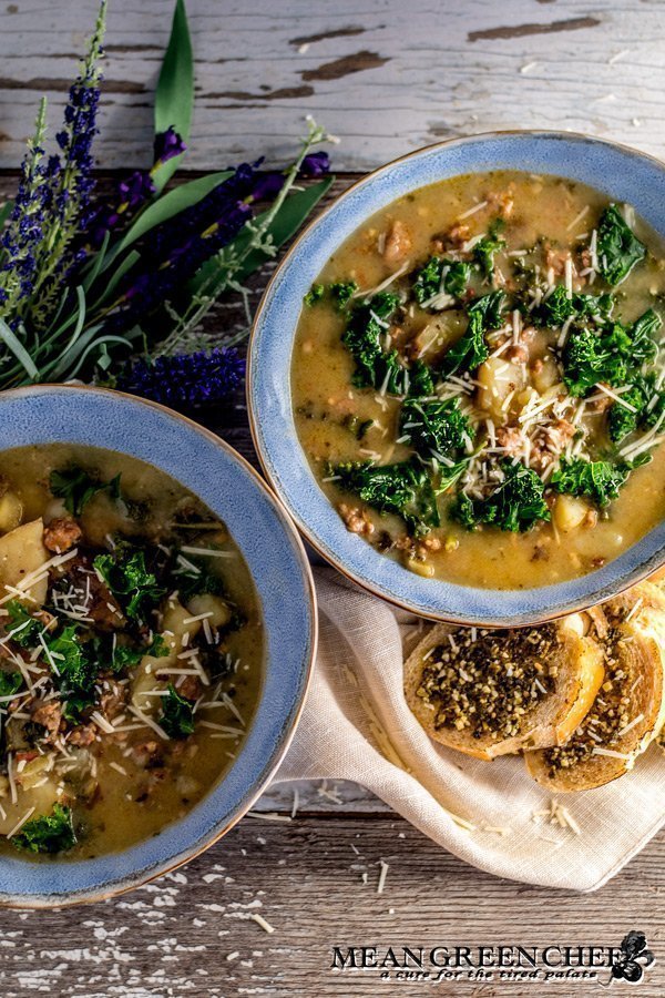 Overhead photo of Zuppa Toscana in two large blue bowls with rough edges. A small beige linen napkin and bread spread with pesto. Mean Green Chef