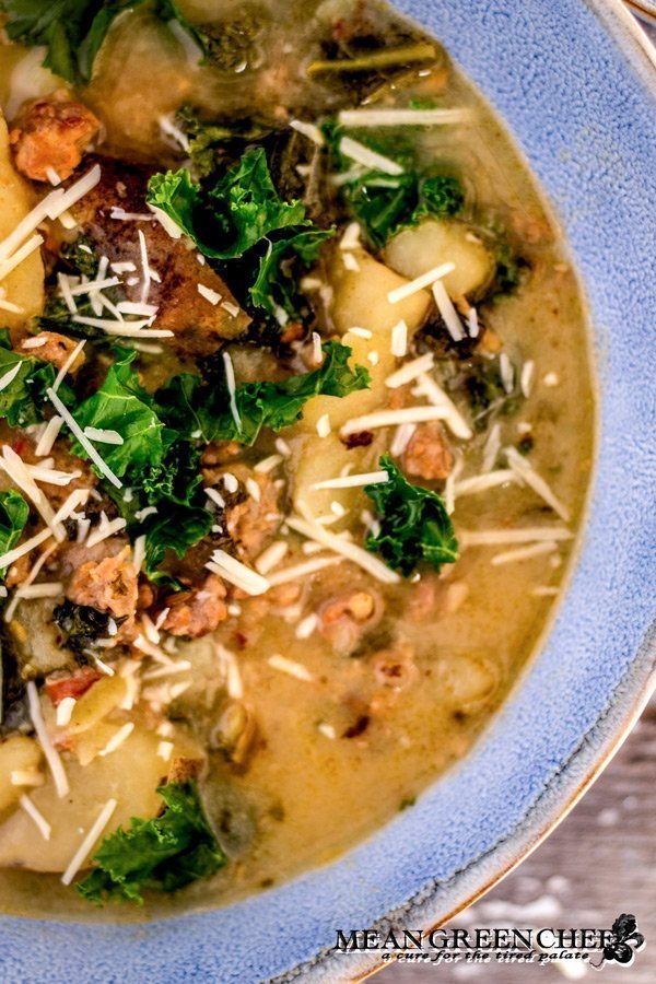 Close up photo of Zuppa Toscana in a large blue bowls with a rough edge. A small beige linen napkin and bread spread with pesto. Mean Green Chef
